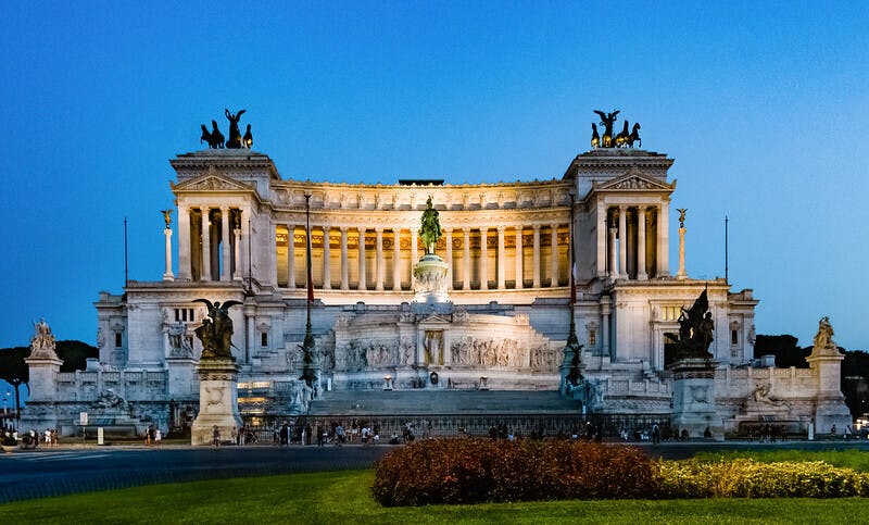 Osvětlená neoklasicistní budova se sloupy a sochami při západu slunce s centrální jezdeckou památkou a zdobenými sochami.