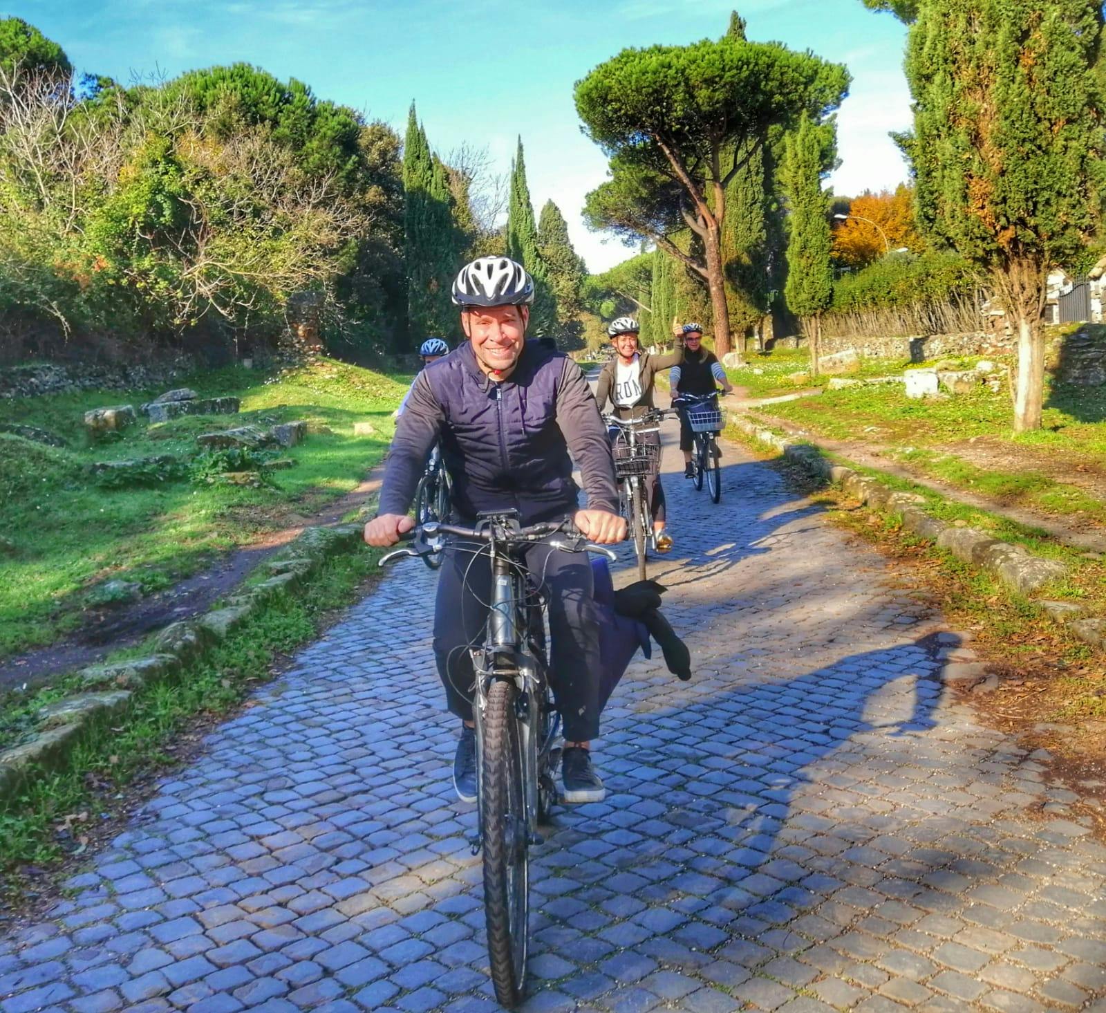 People riding bicycles on a cobblestone path, surrounded by lush greenery and tall trees, under a clear blue sky.