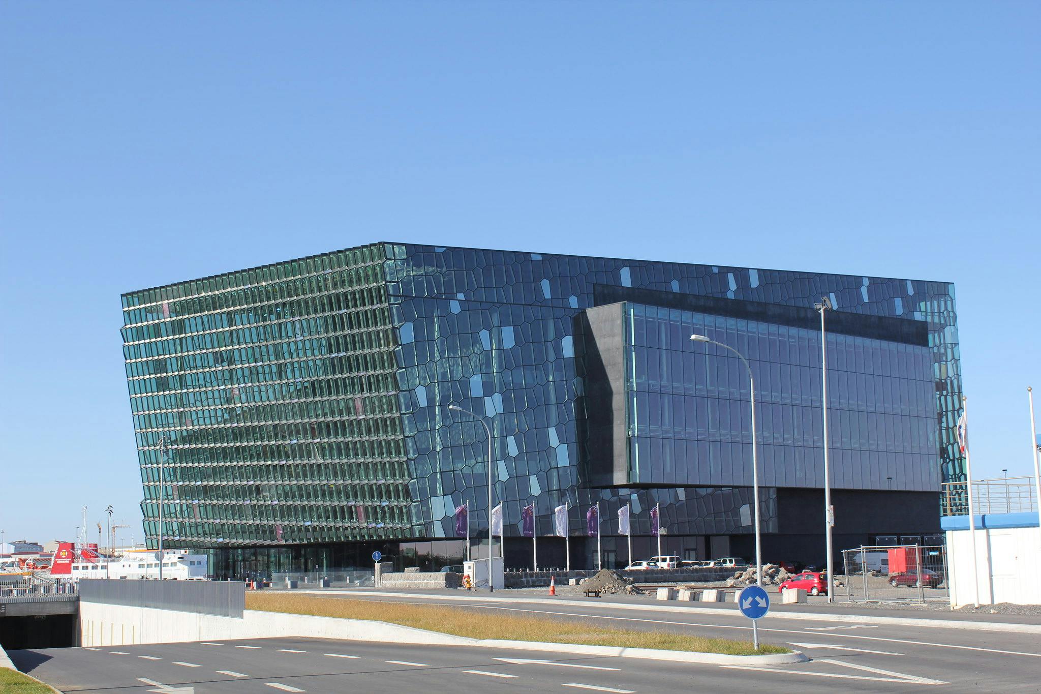 A modern glass and steel building with geometric patterns on the exterior under a clear blue sky, located next to a road.