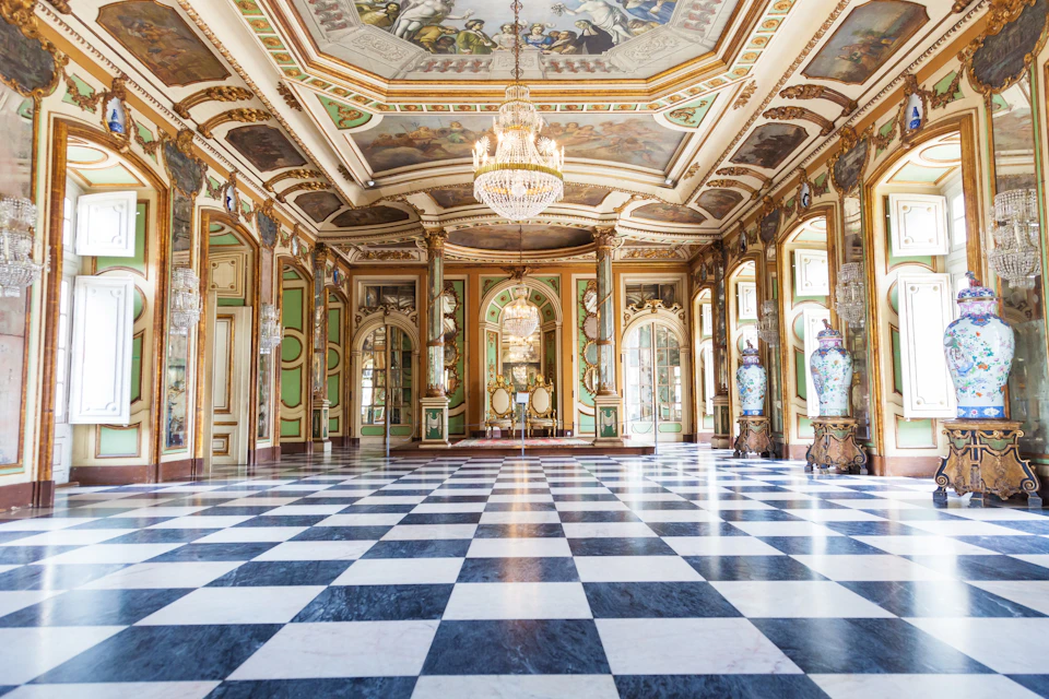 An ornate, empty ballroom with a checkered floor, chandeliers, frescoed ceiling, and large vases along the walls.