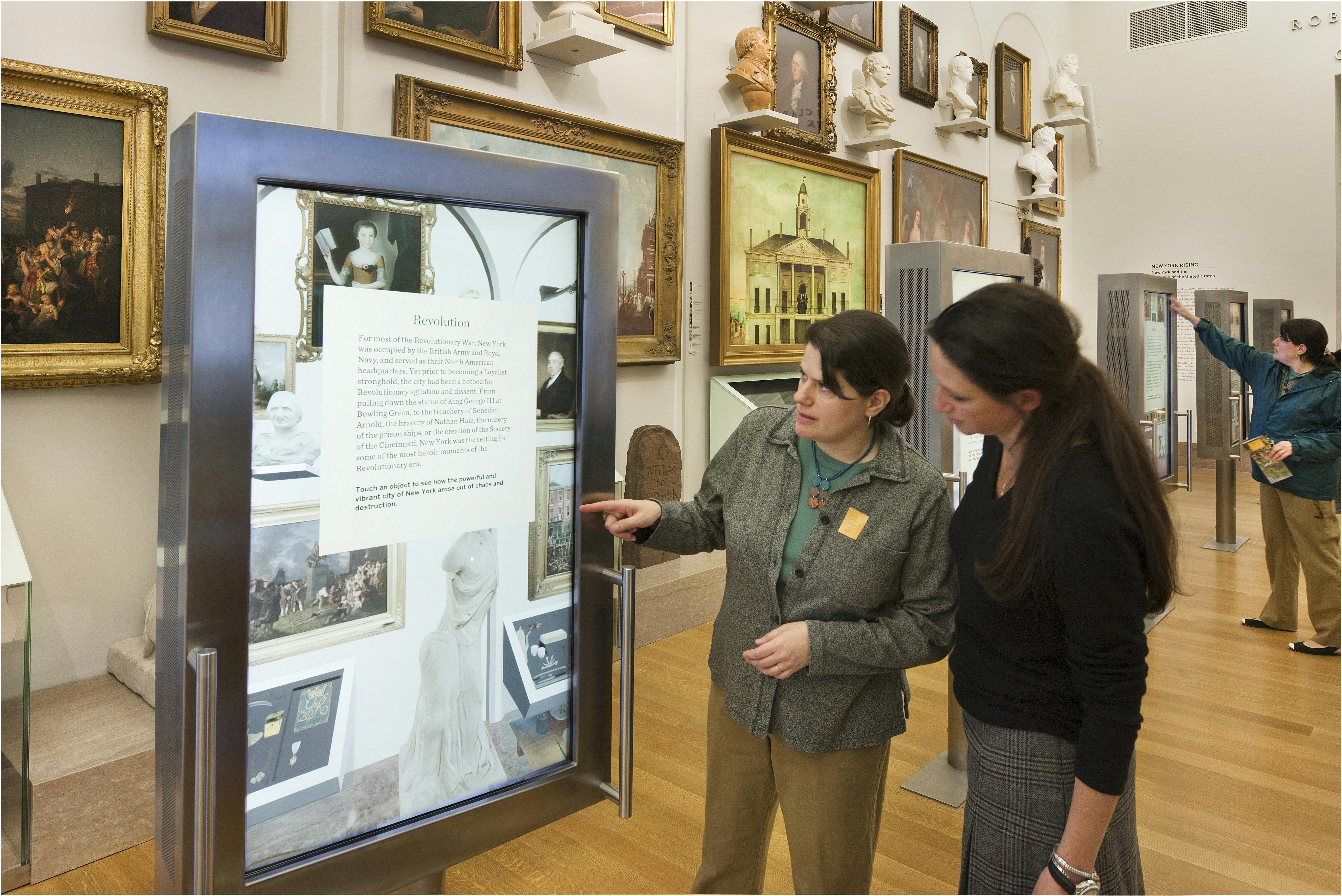Δύο γυναίκες σε ένα μουσείο συζητούν για ένα έκθεμα, με πίνακες και γλυπτά στους τοίχους και έναν άλλο επισκέπτη στο παρασκήνιο.