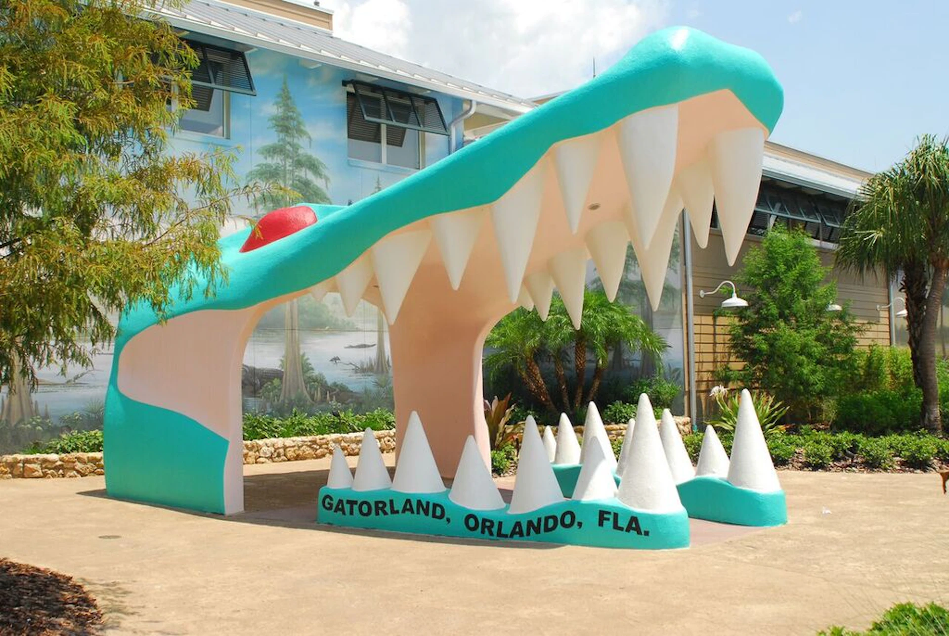 Large alligator mouth entrance with sharp teeth at Gatorland, Orlando, FL, with a mural and plants in the background.