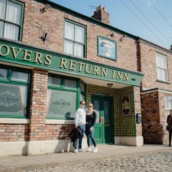 Two people stand in front of the "Rovers Return Inn," a brick building with green trim and large windows. A cobblestone street is in the foreground.