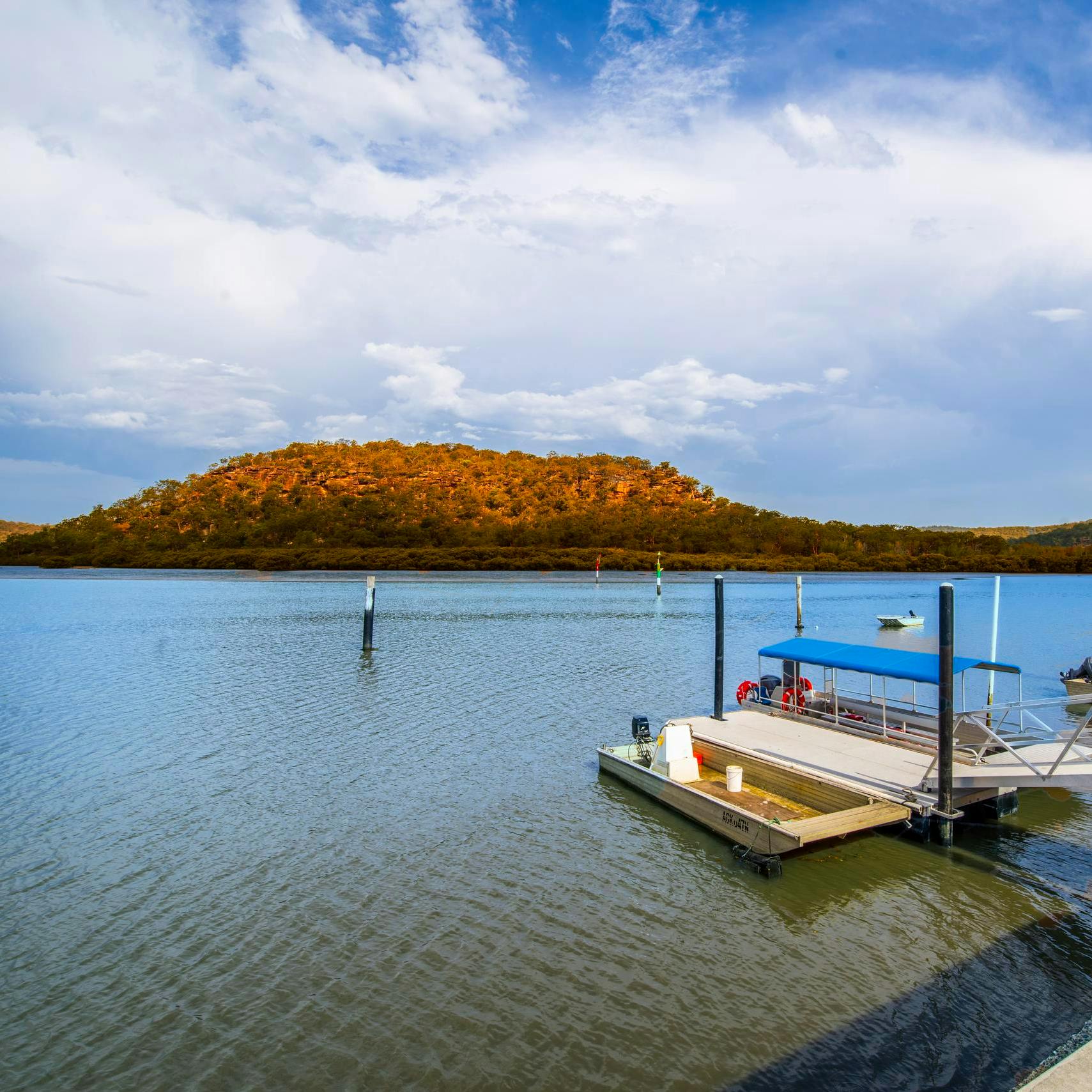 一个码头，一艘蓝色屋顶的船在平静的水面上，背景是一座森林山丘，在一片多云的天空下。