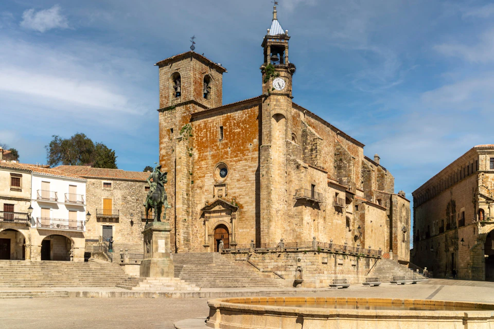 Historic stone church with a clock tower, a horse statue in the square, and stairs leading to the entrance under a blue sky.