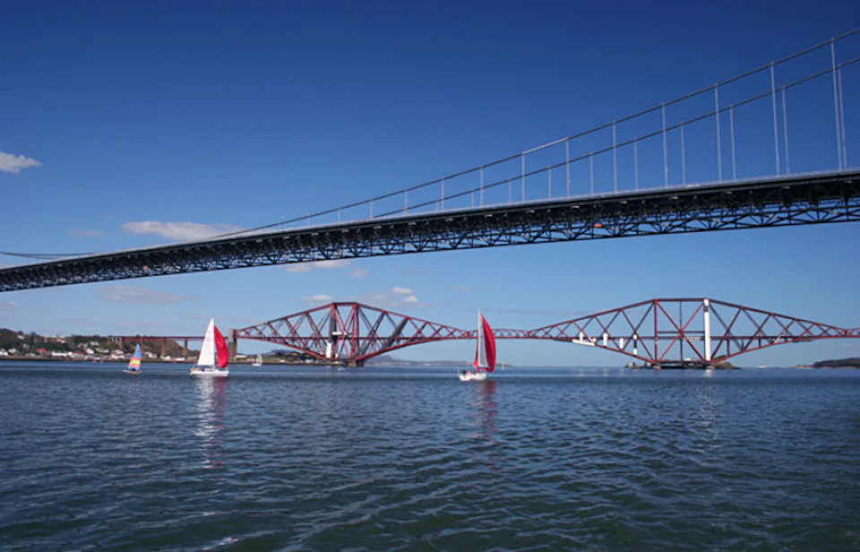 Two bridges crossing over a body of water with three sailboats, each featuring red and white sails, against a clear blue sky.