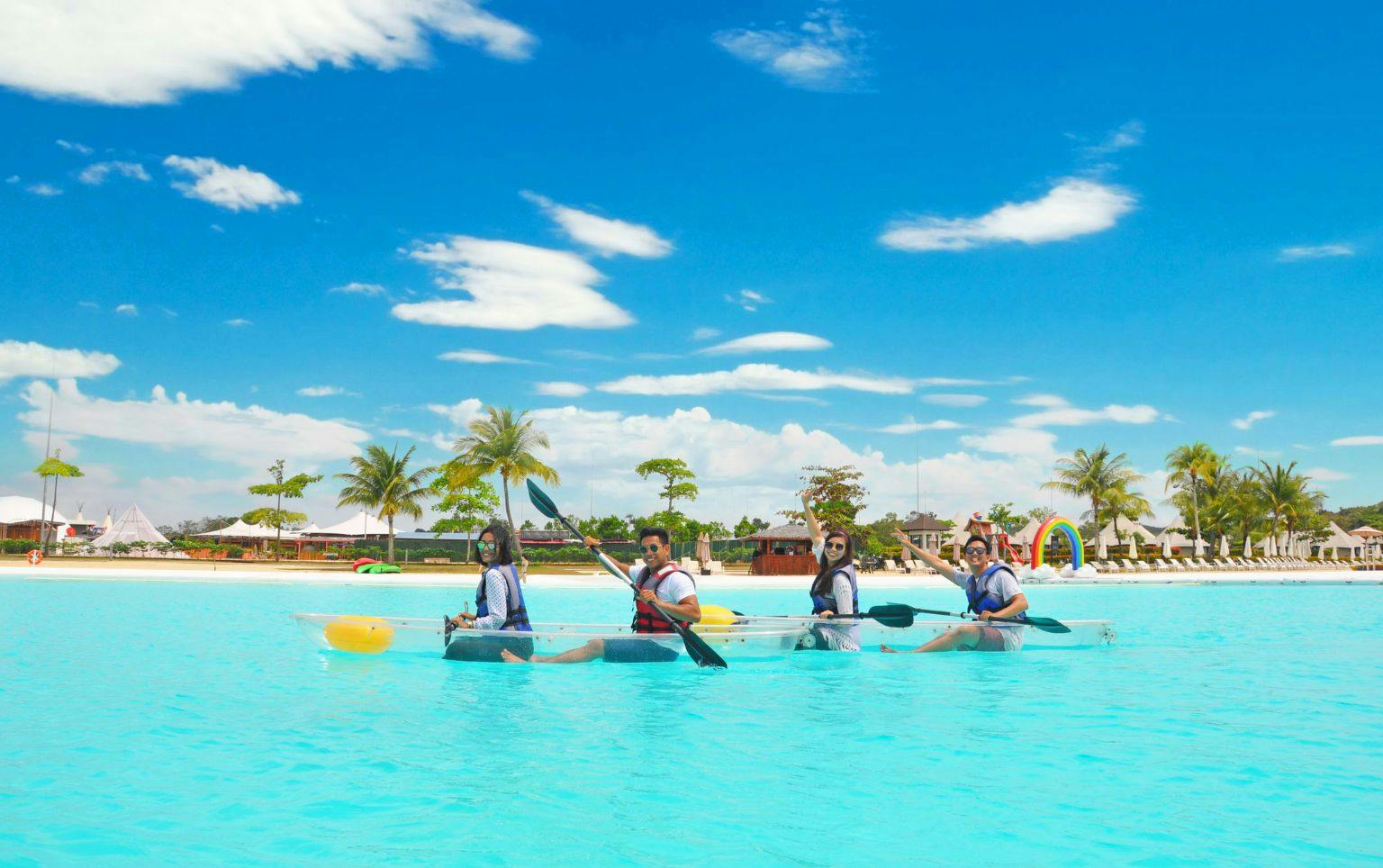 Four people paddle a transparent kayak on a bright blue water lagoon, with palm trees and a clear sky in the background.