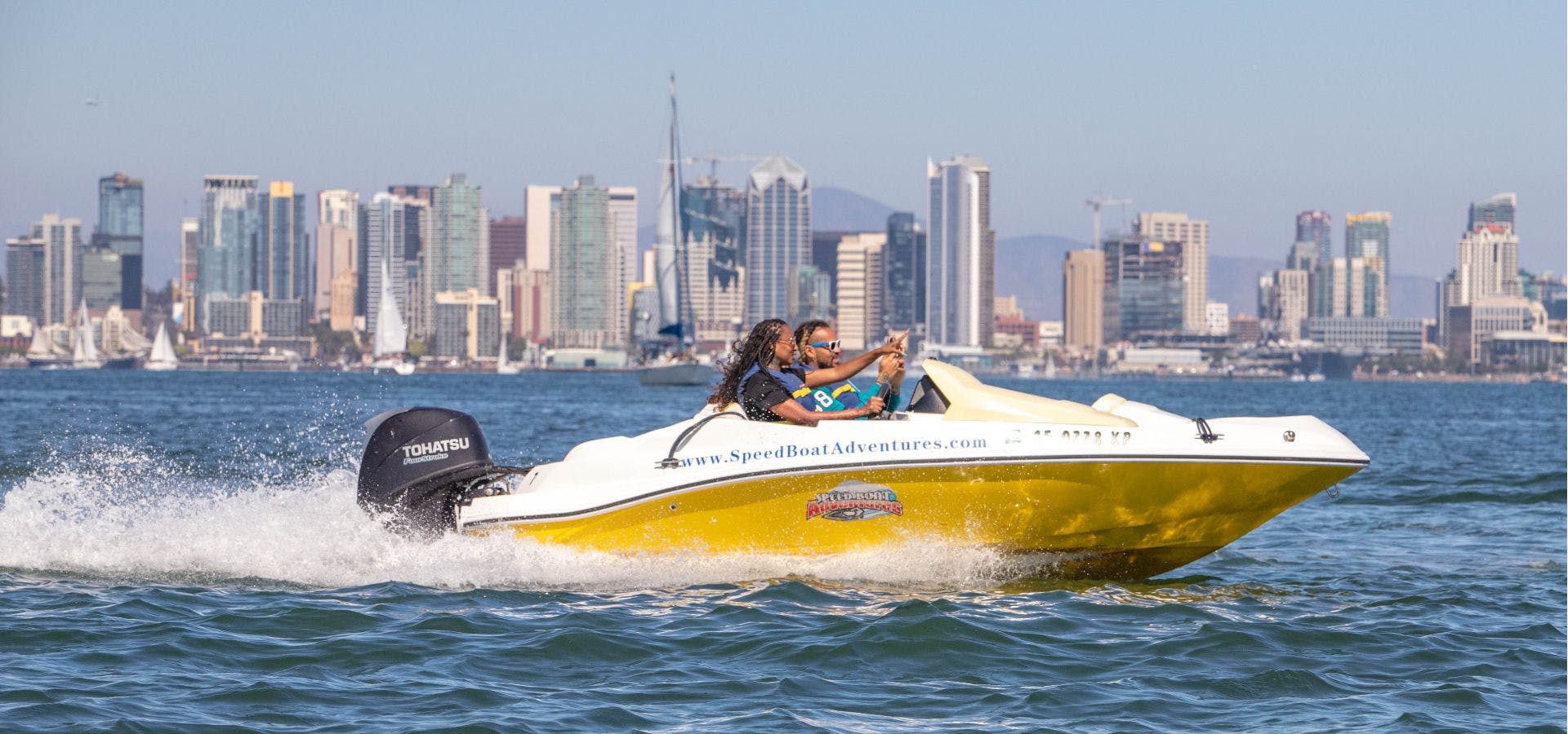 Una lancha rápida amarilla con dos pasajeros navega por el agua frente al horizonte de una ciudad en un día despejado.