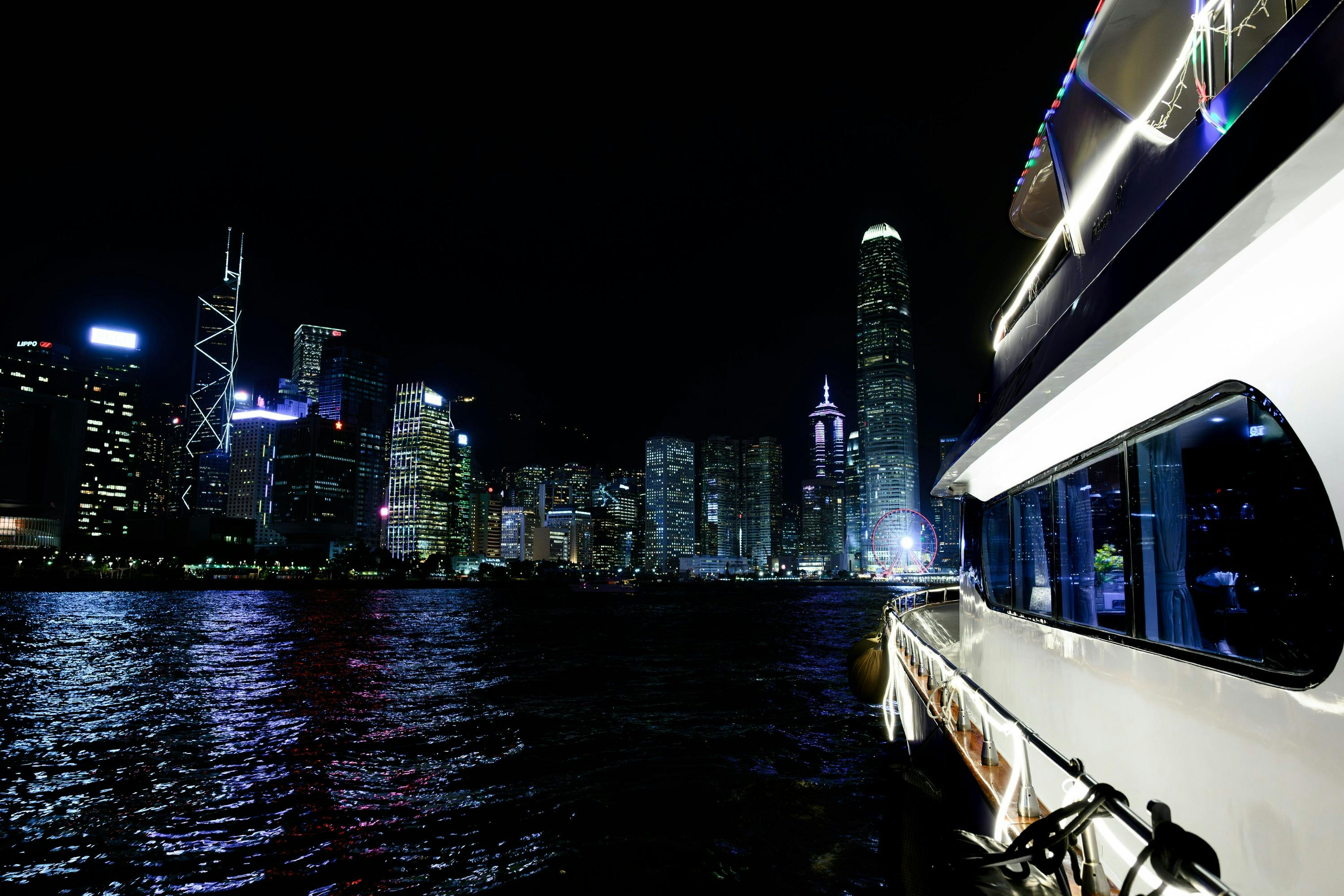 A city skyline at night with illuminated skyscrapers reflected on water, and part of a boat on the right side of the frame.