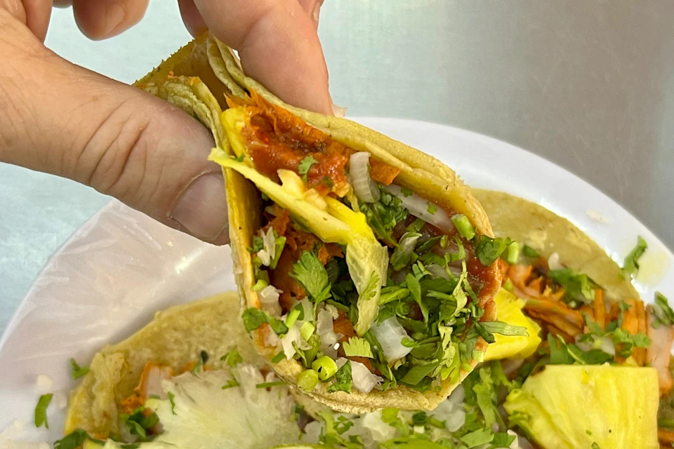 Hand holding a freshly made taco al pastor filled with marinated pork, pineapple, chopped onion, and cilantro.