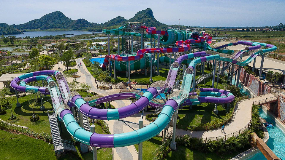 Water park with colorful water slides, looping tubes, and pathways. Mountains and greenery visible in the background.
