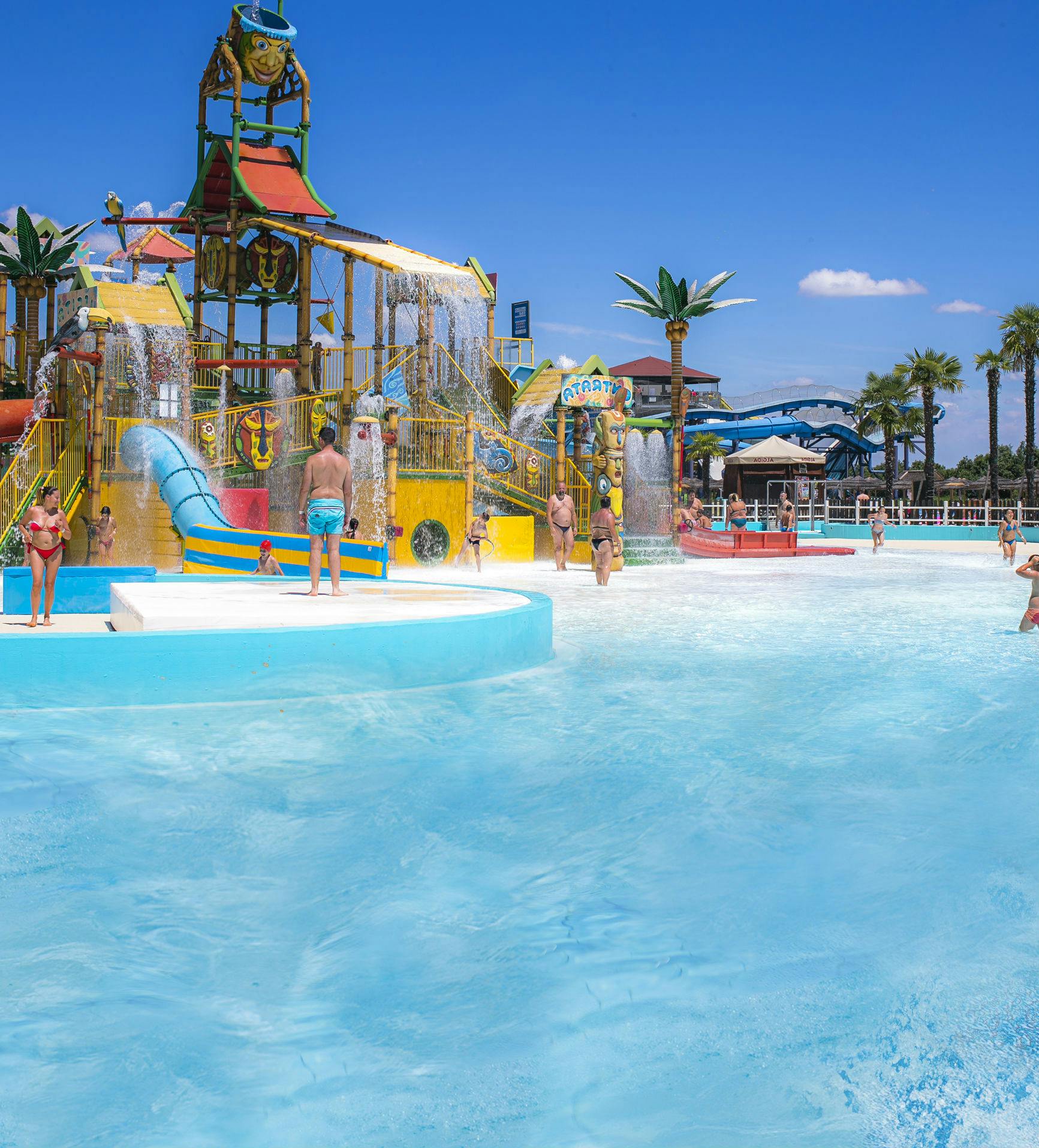 People enjoying a water park with slides, splash structures, and palm trees under a clear blue sky.
