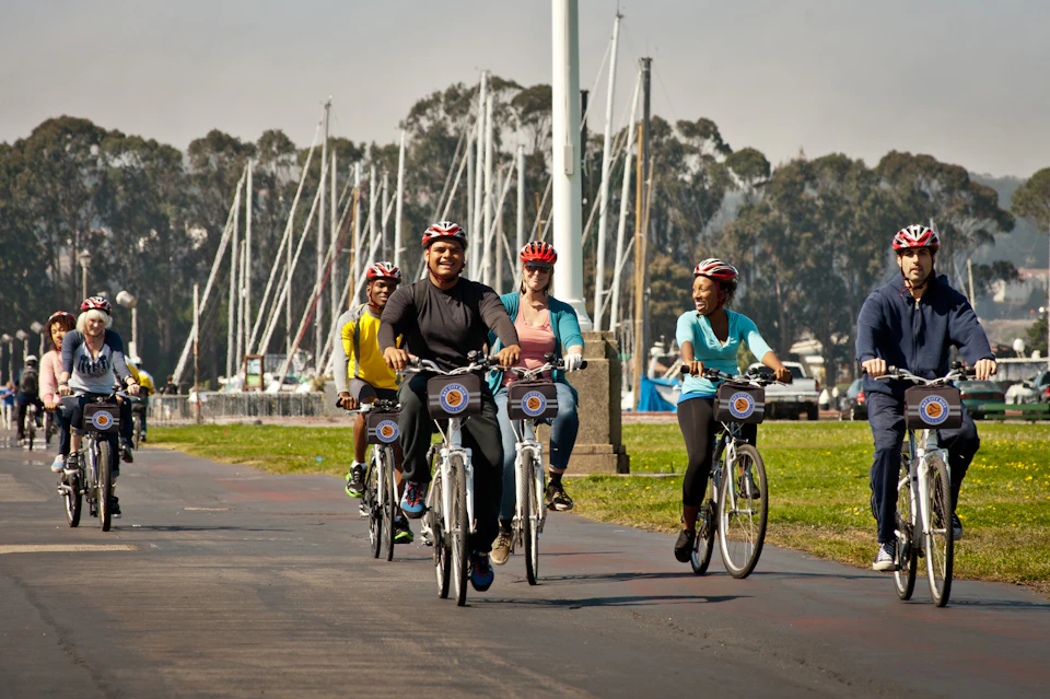 A group of six people riding rental bikes and wearing helmets on a paved path near a marina and green grass.