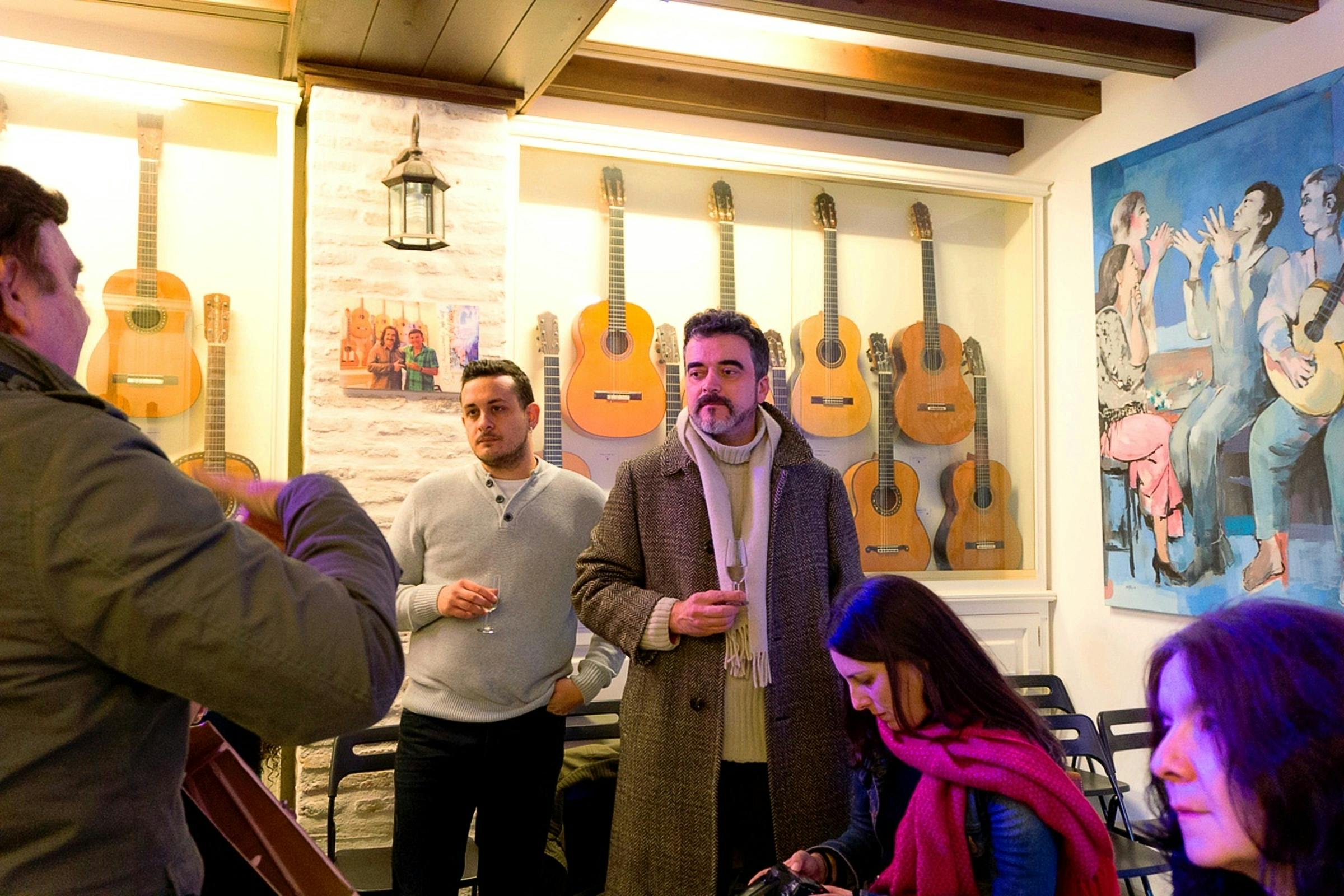 Люди, стоящие и сидящие в комнате с несколькими гитарами на стене и большой картиной на заднем плане.