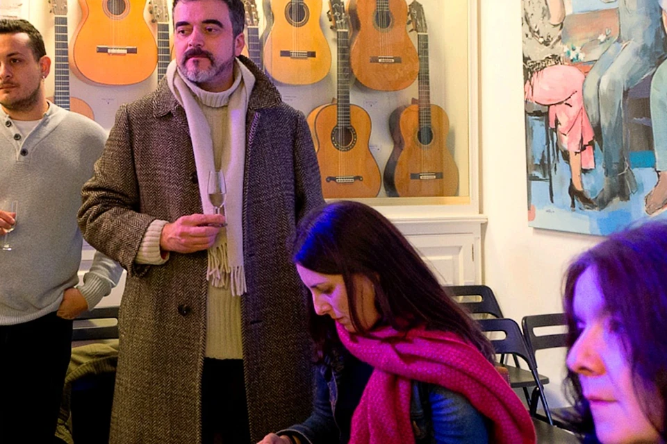 People standing and sitting in a room with several guitars displayed on the wall and a large painting in the background.