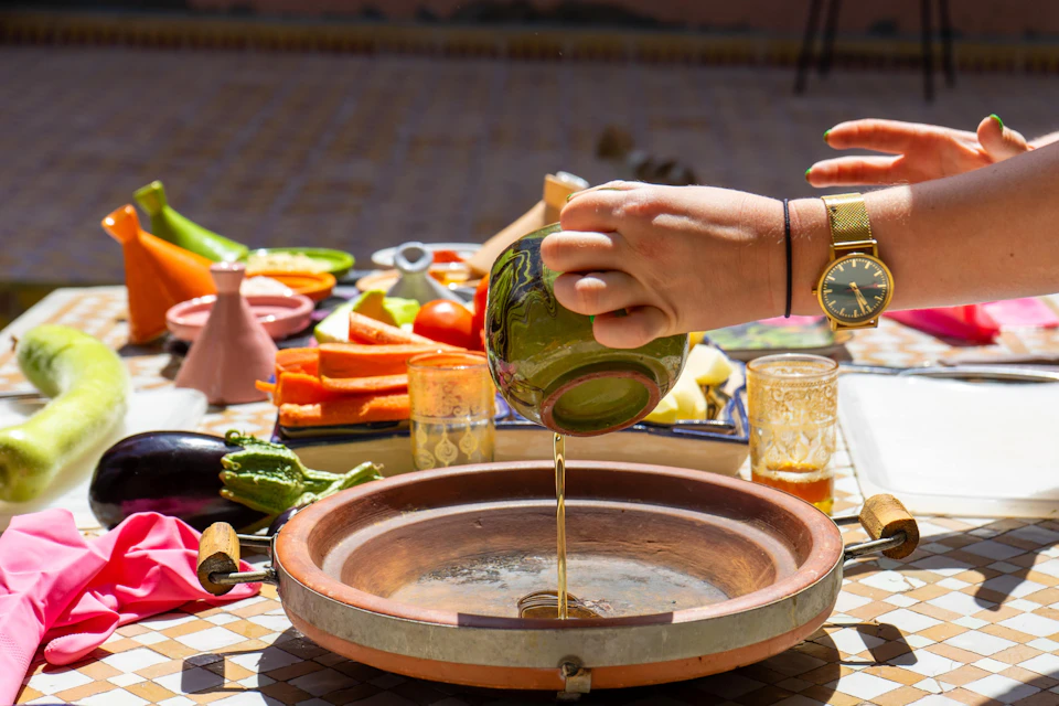 A hand pouring oil into a clay dish on a table set with various cut vegetables and condiments in an outdoor setting.