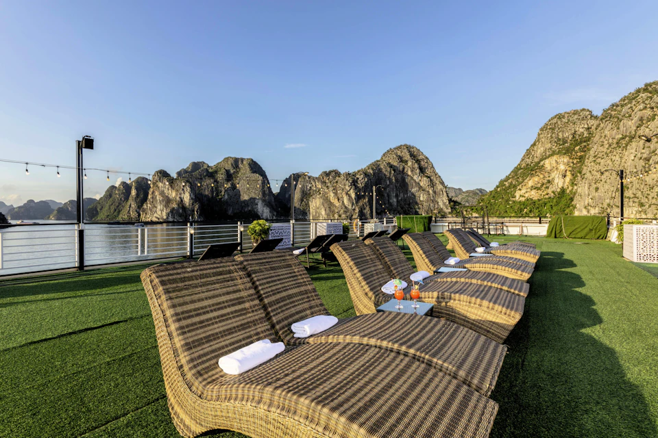 Outdoor lounge chairs with towels and drinks on small tables, overlooking water and rocky hills under a clear sky.