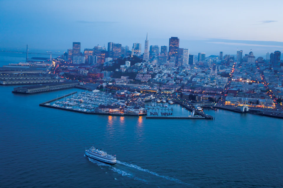 Aerial view of a city skyline at dusk with a boat cruising in the foreground and a marina on the left.
