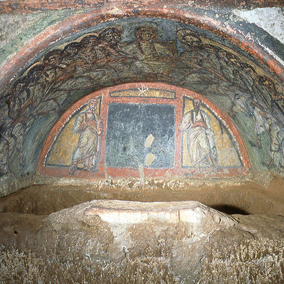 Ancient frescoes depicting religious figures, with a central blue panel, cover a curved ceiling above a stone structure in a cave.