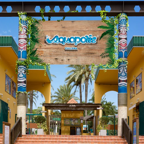 Entré till Aquopolis vattenpark, med kolumner med tiki-tema, palmer och skyltar. Röda tegelsteg leder till biljettområdet.