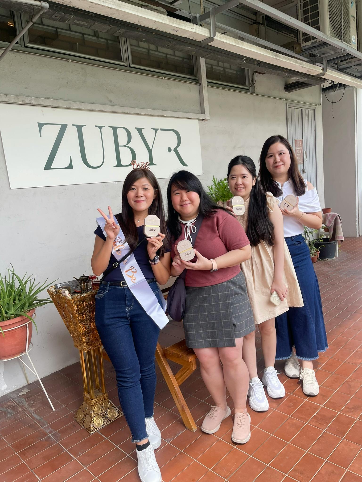 Four women posing together in front of a storefront with the sign "ZUBYR." They are smiling and holding items in their hands.