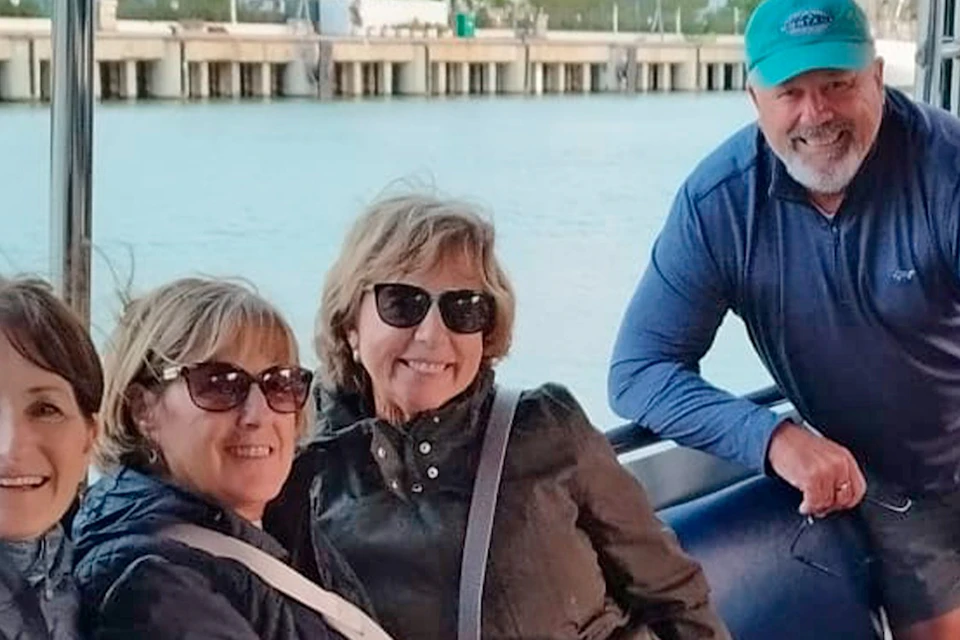 A group of six adults sitting and smiling on a boat with tables, with a waterfront and buildings visible in the background.