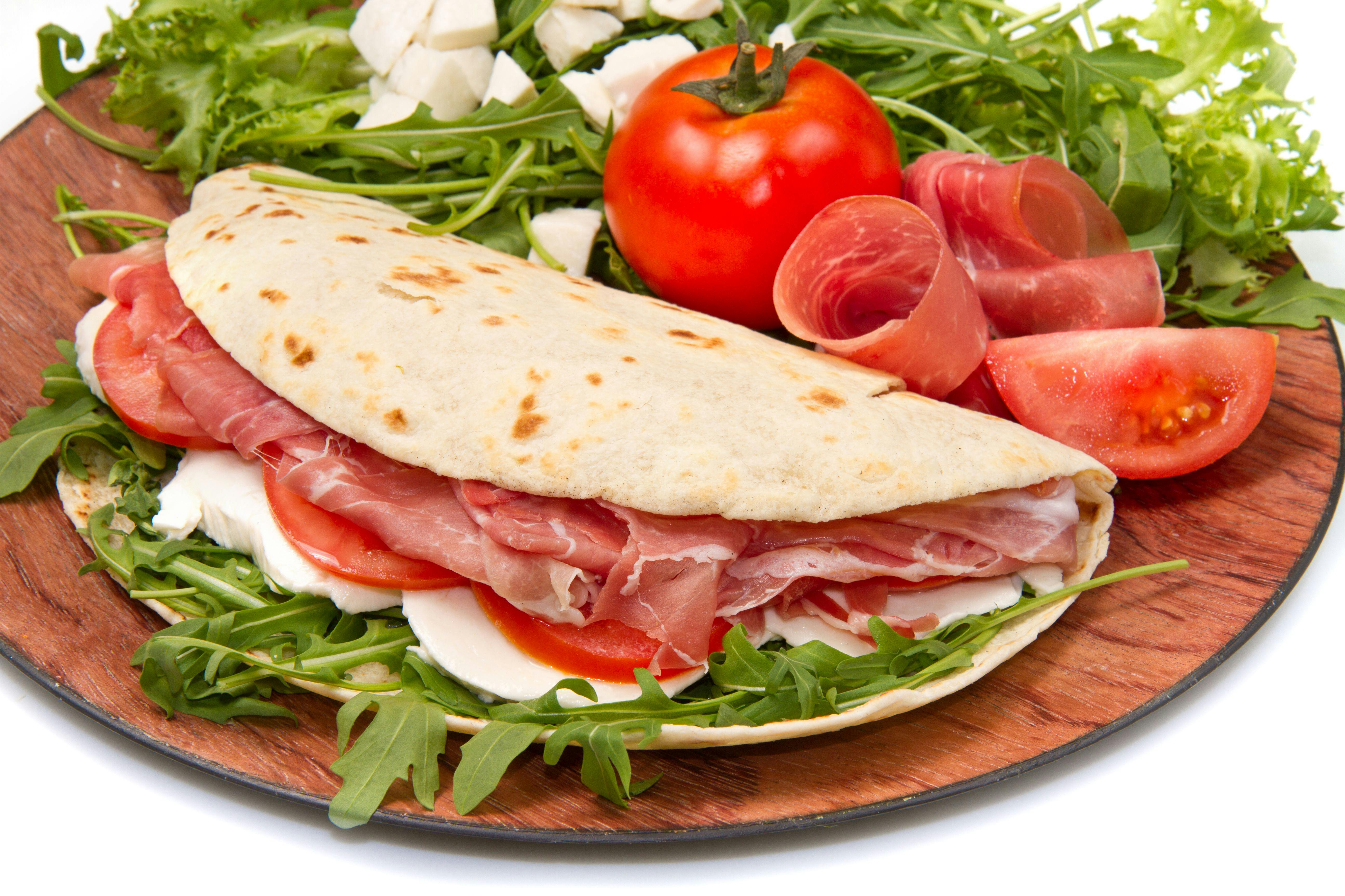 Flatbread sandwich with prosciutto, mozzarella, and tomatoes on arugula, garnished with a whole tomato and mozzarella pieces.
