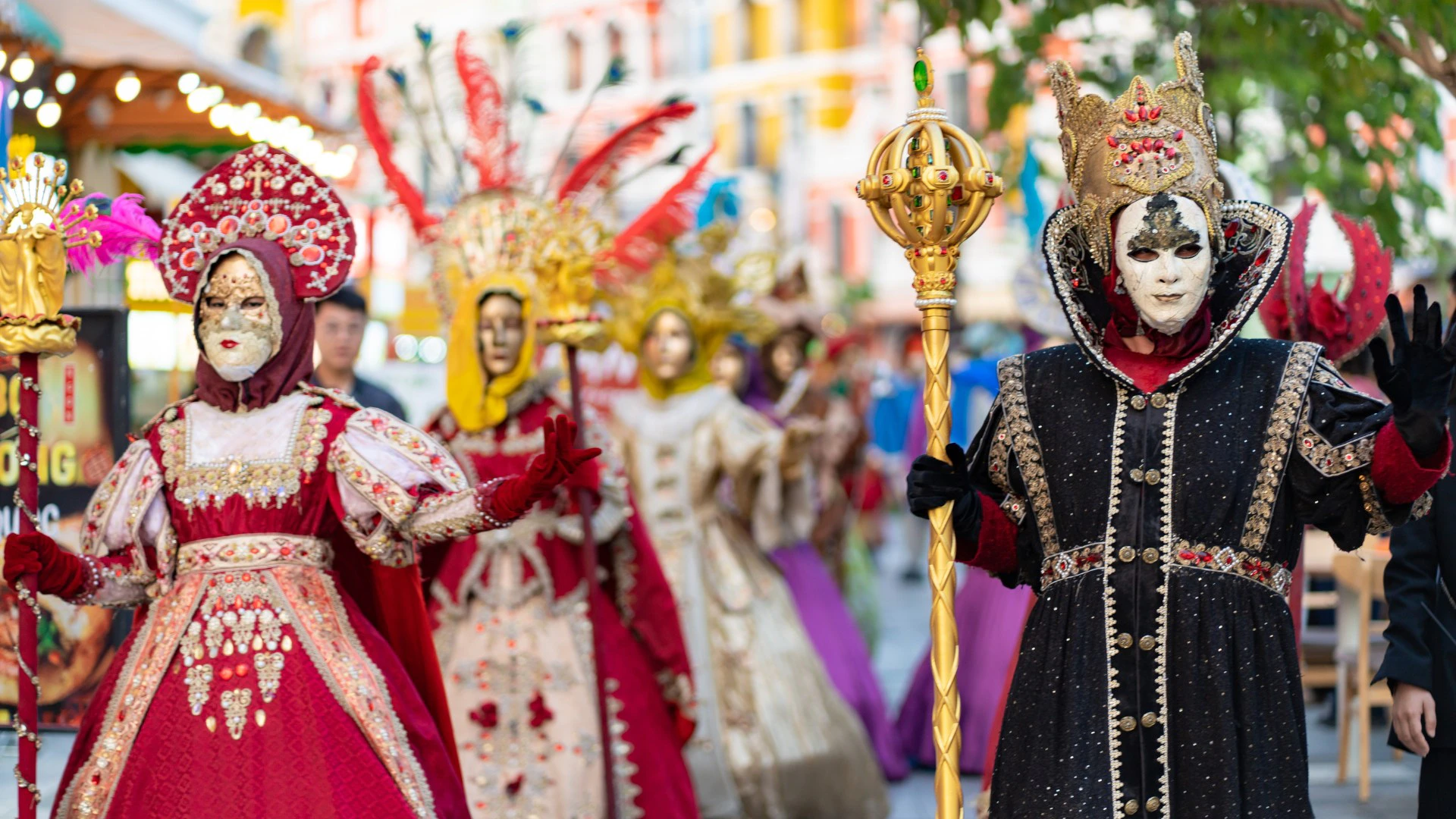 People in ornate, colorful costumes and masks participate in a festive parade on a lively street.
