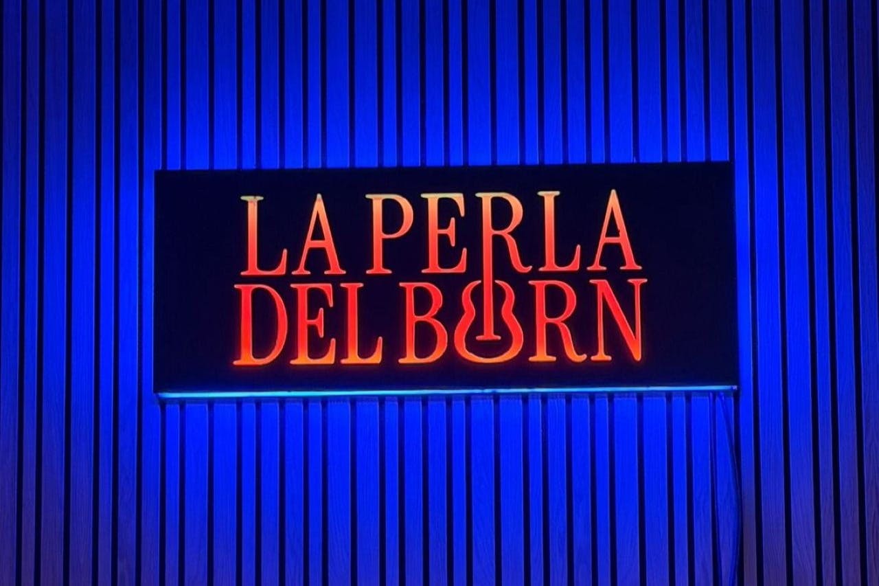 在蓝色背景下，橙红色字母带有吉他形状的带照明标志，上面写着“La Perla Del Born”。
