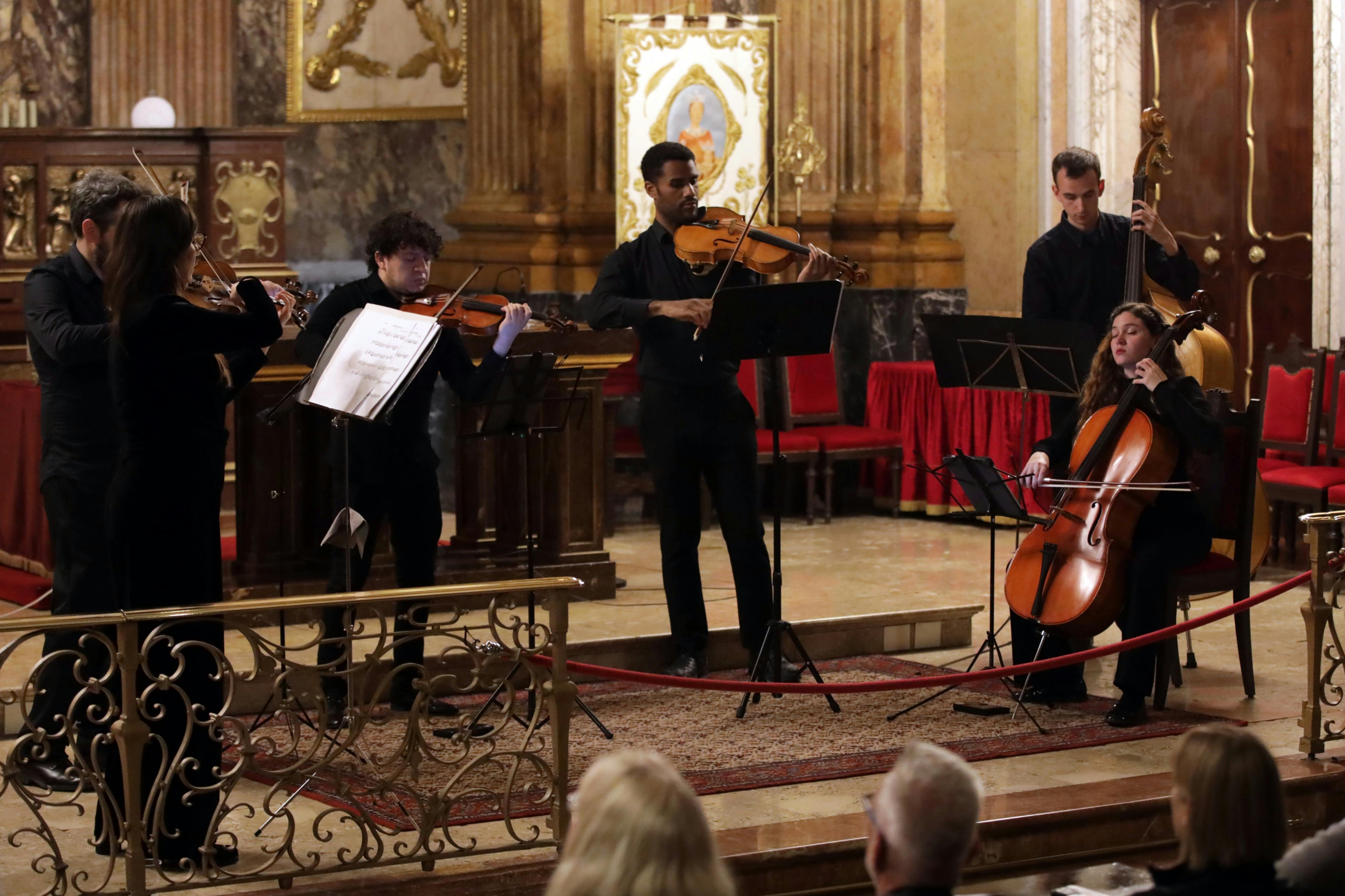 A string quartet performs in a formal setting, with violin, cello, and double bass players visible, observed by an audience.