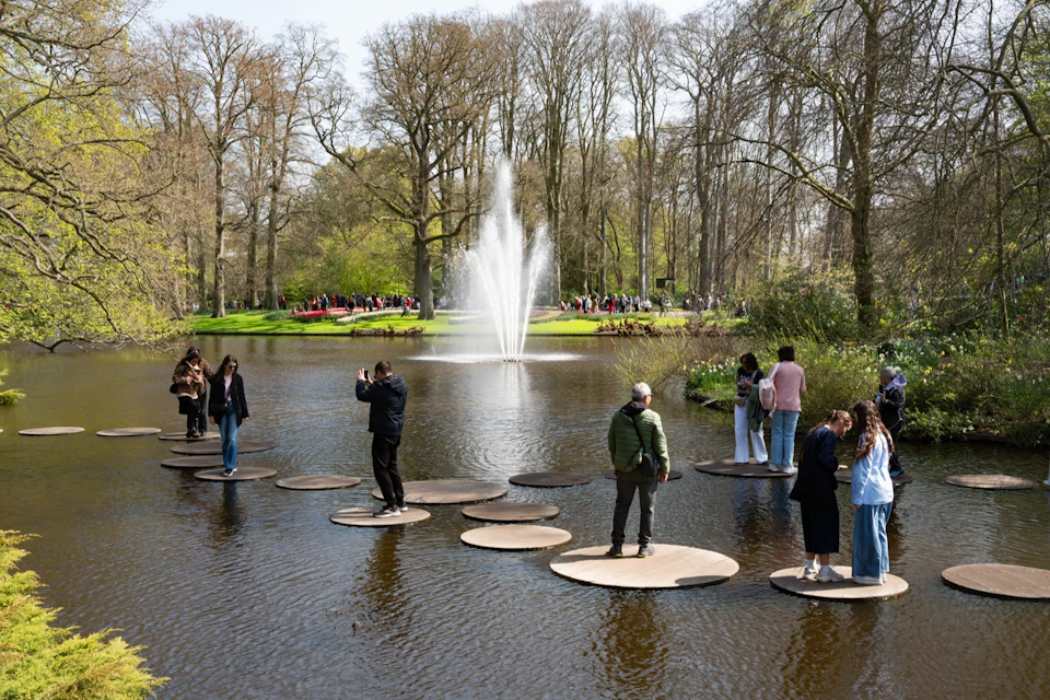 People walking on stepping stones in a pond with a fountain in the center, surrounded by trees and greenery on a sunny day.