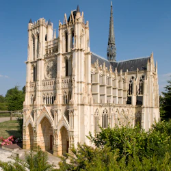 Utsmyckad katedral med höga spiror och välvda ingångar, omgiven av grönska, under en klarblå himmel.
