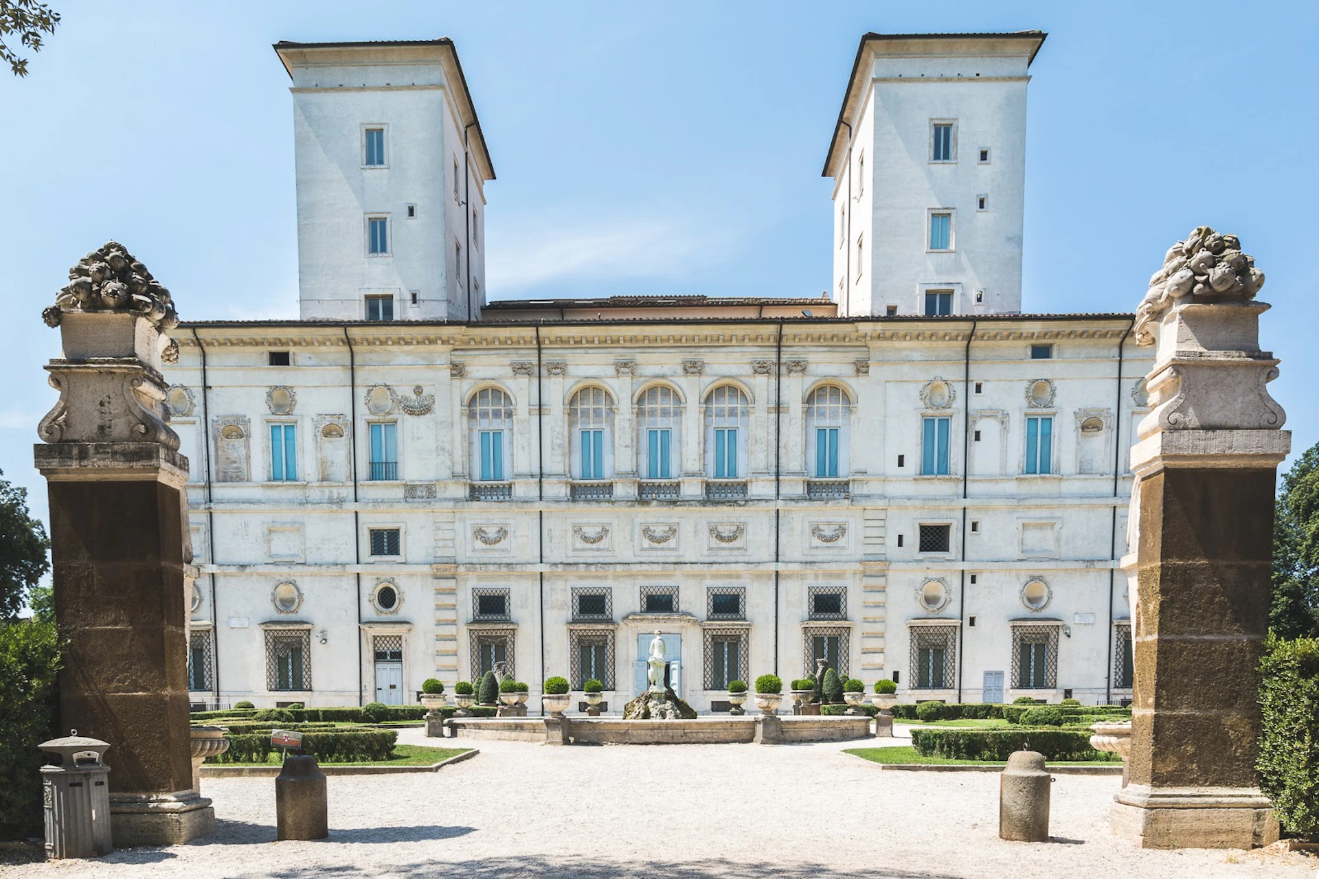 A grand historic building with two tall towers, ornate windows, a central statue, and manicured gardens under a clear blue sky.