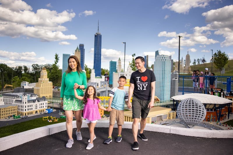 A smiling family of four walks hand-in-hand in front of a miniature cityscape under a bright, partly cloudy sky.