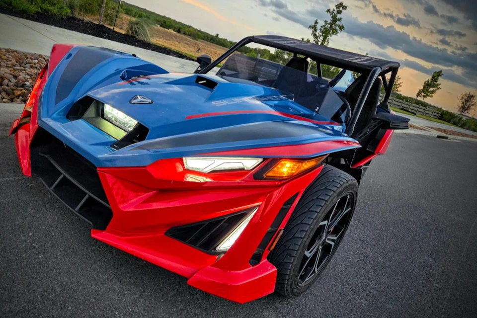 A sporty three-wheeled vehicle with a blue and red design parked on a paved road, with trees and sky in the background.