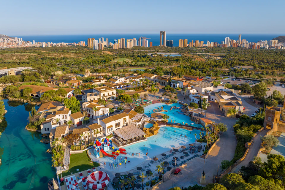 Aerial view of a resort with pools, slides, and buildings surrounded by greenery, with a city skyline and sea in the background.
