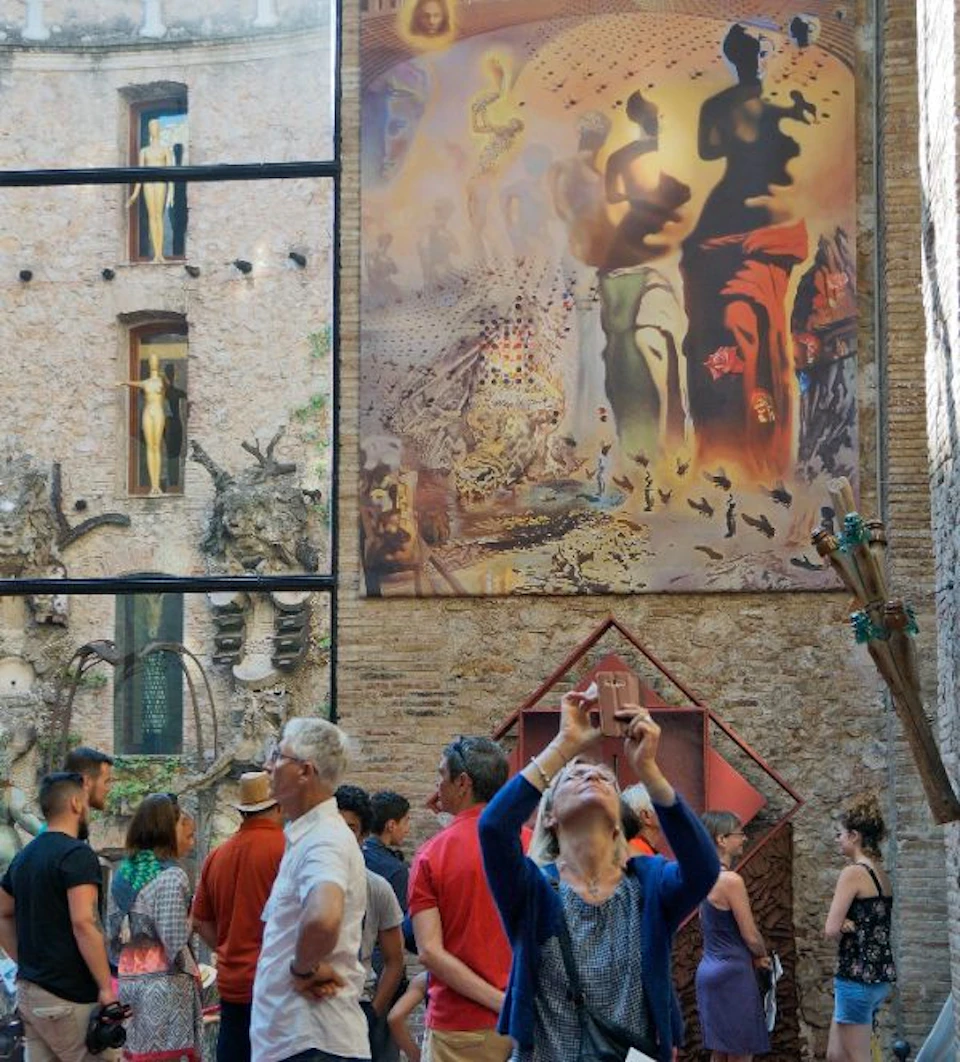 A crowd of people stands in front of a building with surrealist art, while one person takes a photo with a smartphone.