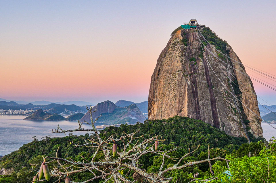 A large granite rock formation with a building on top, surrounded by dense green vegetation and distant mountains under a colorful sky.