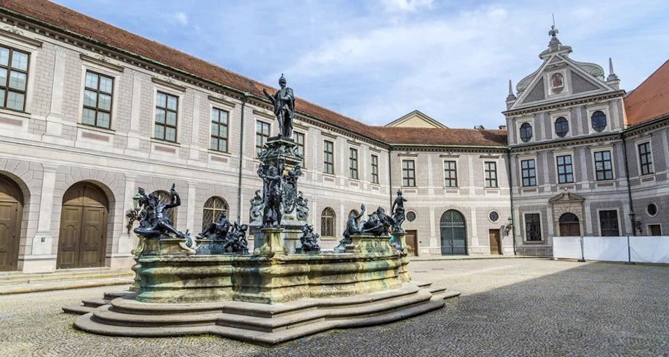 Ornate fountain with multiple statues in a paved courtyard, surrounded by a large historic building with arched windows and red-tiled roof.