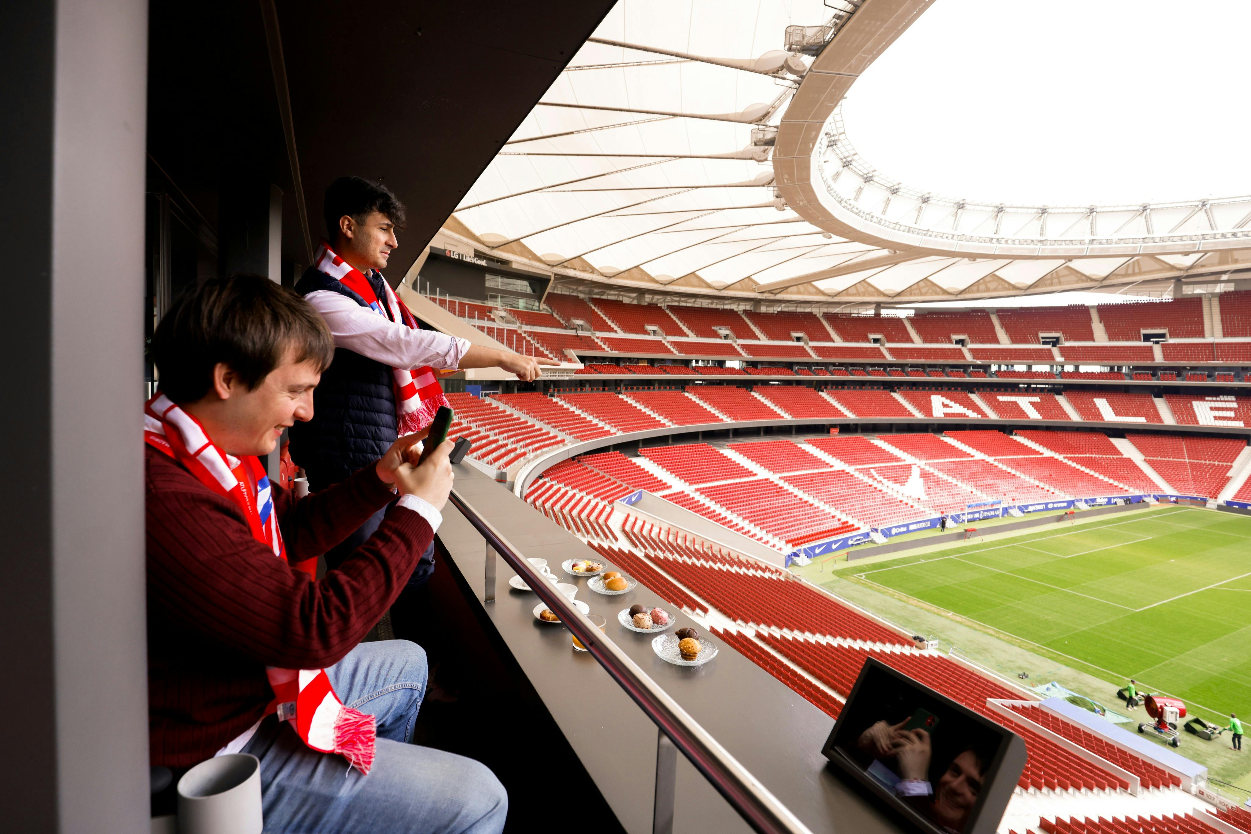 Δύο άνδρες που φορούν κασκόλ απολαμβάνουν τη θέα του σταδίου από ένα ιδιωτικό κουτί. Ο ένας τραβάει μια φωτογραφία και το φαγητό τοποθετείται στο κιγκλίδωμα.