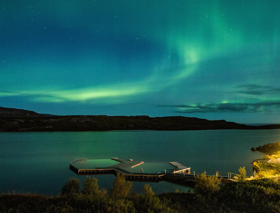 A serene lake at night with a wooden dock extending into the water and the Northern Lights illuminating the sky above.