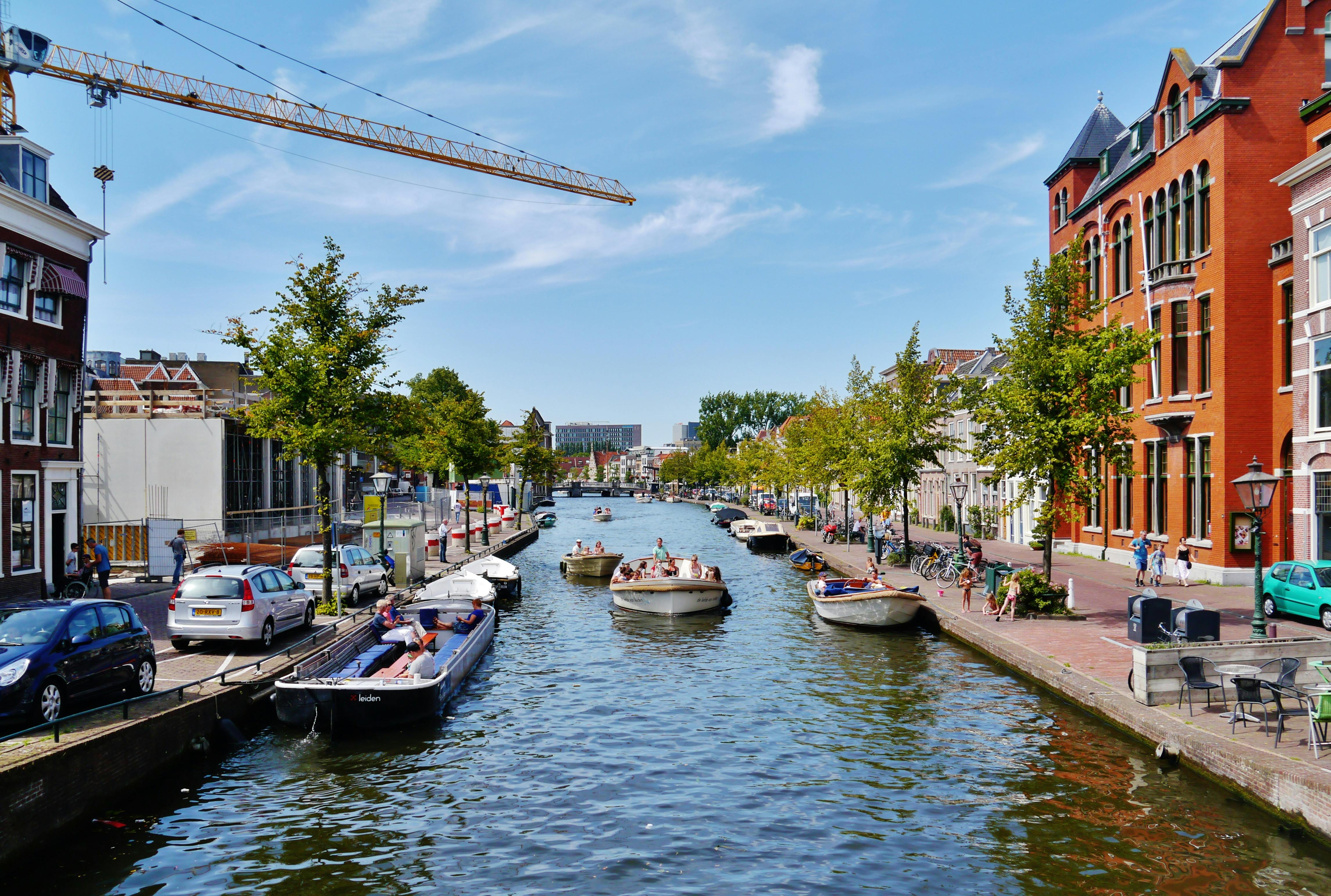 Za slunečného dne si lidé odpočinou na lodích plujících po kanále lemovaném stromy, zaparkovanými auty a budovami s jeřábem nad hlavou.