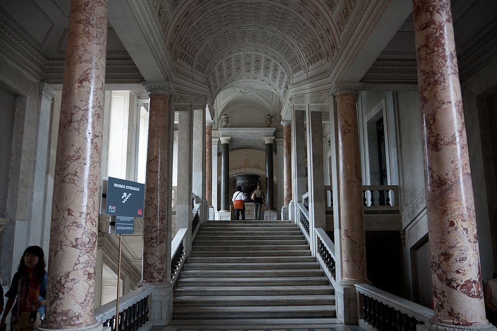 Grand staircase with pink marble columns, arched ceiling, and a sign for Museo Etrusco. Two people stand at the top.