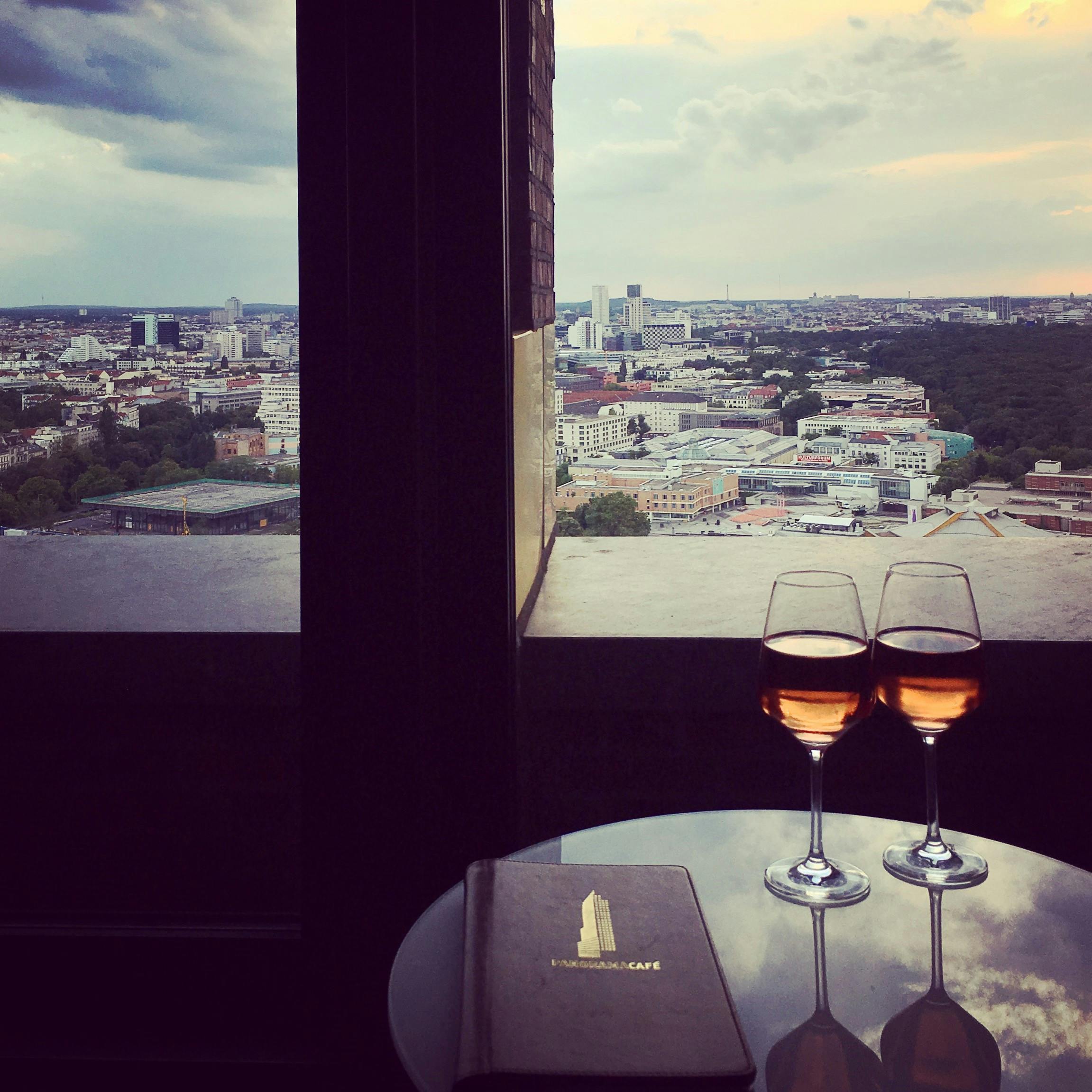 Twee glazen wijn en een menukaart op een tafel met uitzicht op een stadsgezicht in de schemering door een groot raam.