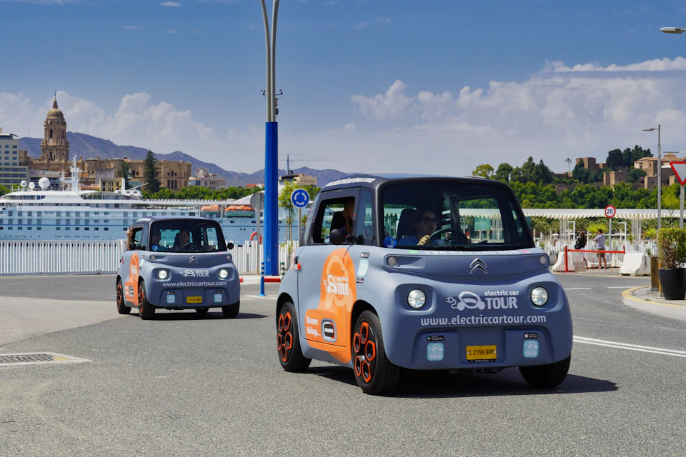 Two small electric cars with "electric car tour" branding drive along a sunny street with mountains and buildings in the background.