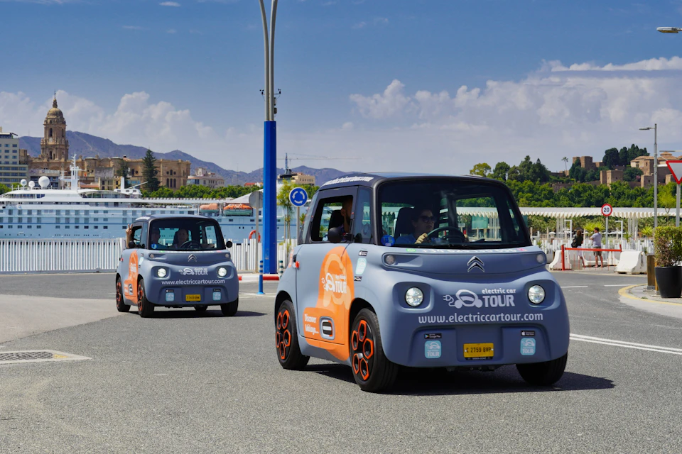 Two small electric cars with "electric car tour" branding drive along a sunny street with mountains and buildings in the background.