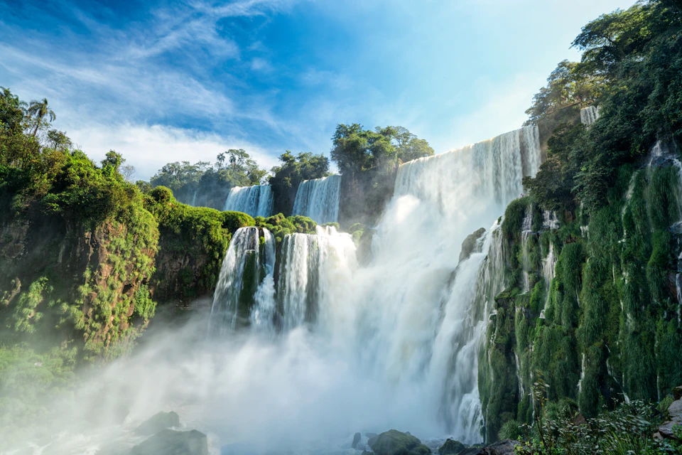 Multiple roaring waterfalls cascade down lush green cliffs under a clear blue sky. Dense mist rises from the base.