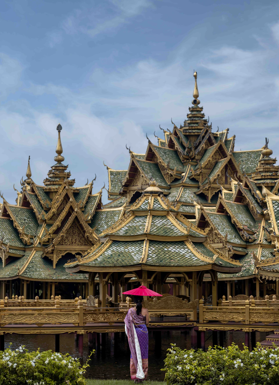 A person holding a red umbrella stands in front of a detailed, ornate, gold and green-roofed temple complex under a cloudy sky.