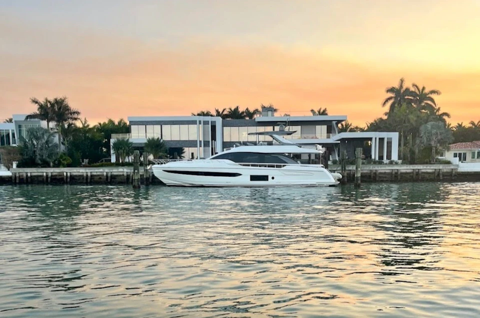 A white yacht docked in front of a modern waterfront house during a pastel-colored sunset.