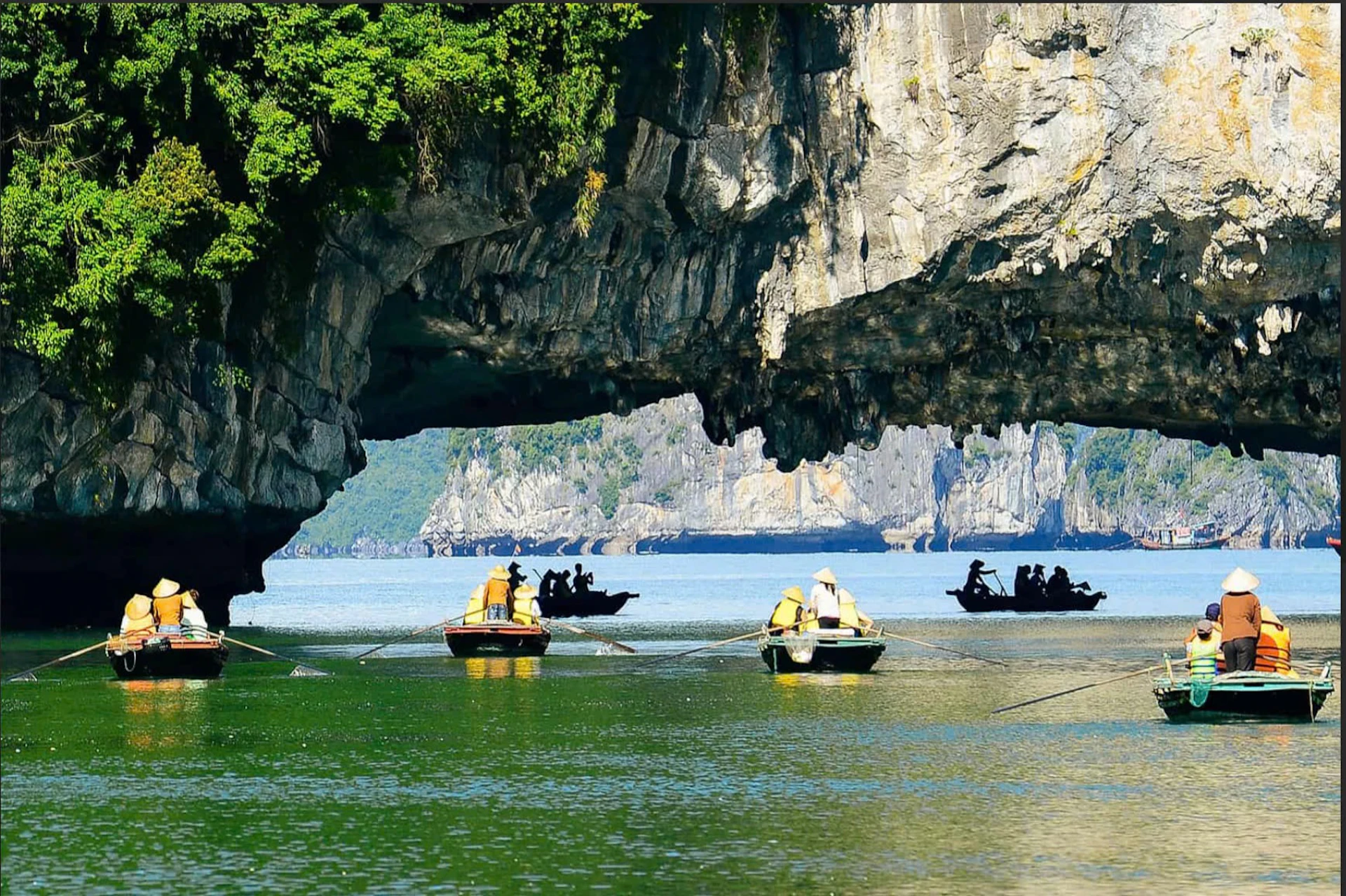 Small boats with people rowing under a large rock arch on a greenish-blue body of water, with cliffs in the background.